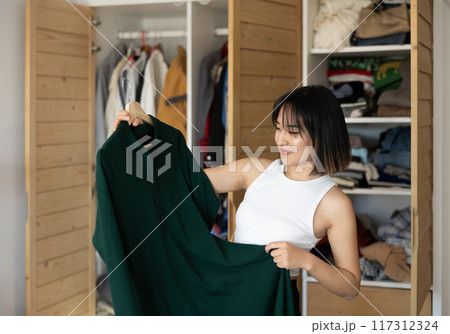 Happy young Asian girl trying to choose a dress standing by wardrobe Happy young Asian girl trying to choose a dress standing by wardrobe 117312324