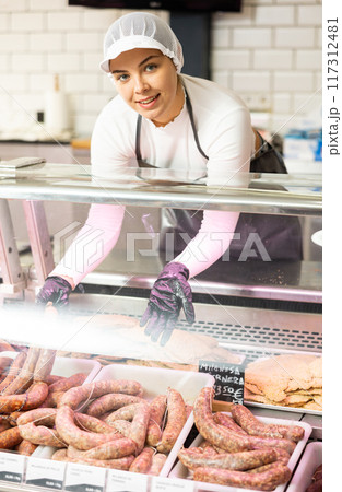 Young female butcher arranging raw sausages in glass counter Young female butcher arranging raw sausages in glass counter 117312481