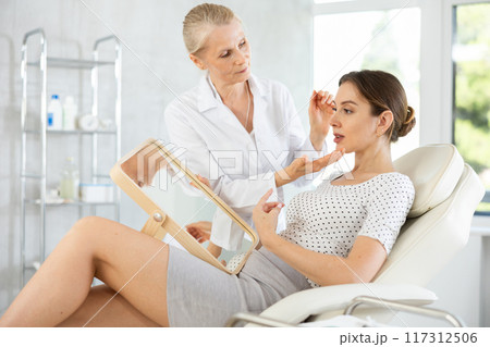 Female patient looking at mirror while professional cosmetologist explaining facial procedure Female patient looking at mirror while professional cosmetologist explaining facial procedure 117312506