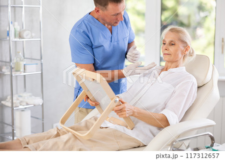 Mature woman sitting with mirror and discussing with male doctor for rejuvenation procedure 117312657
