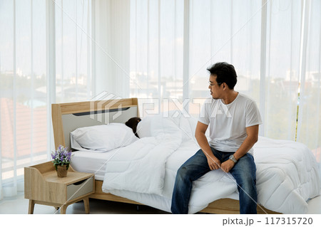 Man sitting on edge of bed in bright bedroom, wearing white t-shirt and jeans, looking to side thoughtfully. woman is sleeping under white blanket, with nightstand and vase of flowers beside bed Man sitting on edge of bed in bright bedroom, wearing white t-shirt and jeans, looking to side thoughtfully. woman is sleeping under white blanket, with nightstand and vase of flowers beside bed 117315720