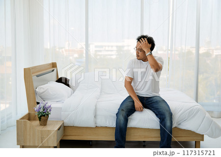 Man sitting on the edge of bed in bright bedroom, wearing white t-shirt and jeans, holding his head as if in thought. woman is sleeping under white blanket beside him, with nightstand and vase of 117315721