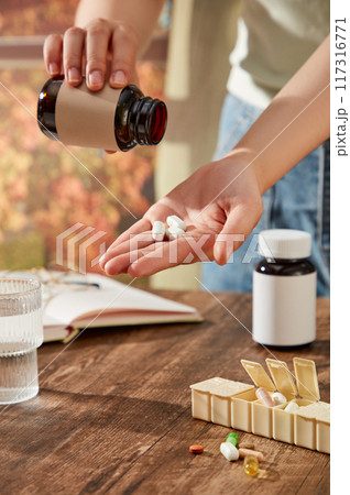 Scene with lifestyle drug advertisement. A woman's hand is pouring medicine in preparation for drinking. Medicine bottles without label on wooden table with glass water cup 117316771