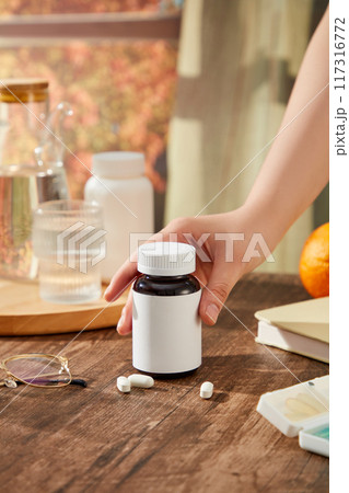 On the wooden table, trays containing water bottles, water glasses and medicine boxes are decorated. hand of woman is preparing to pick up a bottle of medicine unlabeled. Scene for product advertising 117316772