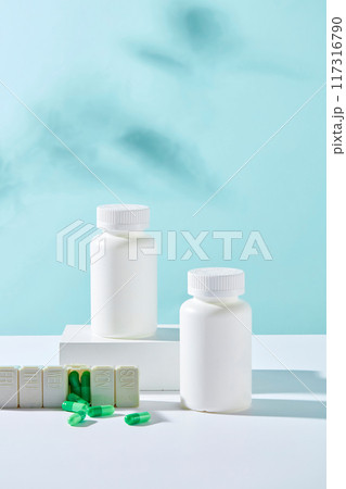 Front view of two white bottles unlabeled displayed on white podiums with medicine daily box and green capsules on blue background. Minimal style for advertising medicine product 117316790