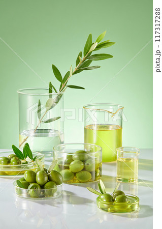 Laboratory concept with beaker and petri dishes filled with olives and olive oil on the green background. Scene for advertising. The bark, leaves and oil of olive peel all have many benefits 117317288