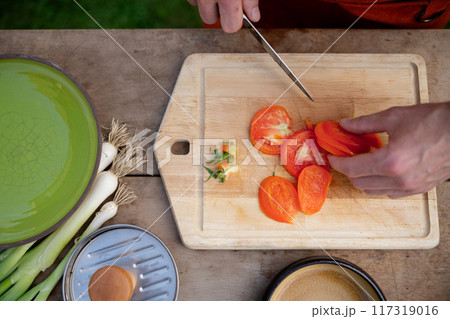 Close up of man holding sharp knife, slicing juicy red tomato. Preparing vegetables for an outdoor barbecue. Close up of man holding sharp knife, slicing juicy red tomato. Preparing vegetables for an outdoor barbecue. 117319016