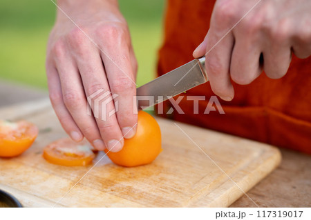 Close up of man holding sharp knife, slicing juicy red tomato. Preparing vegetables for an outdoor barbecue. Close up of man holding sharp knife, slicing juicy red tomato. Preparing vegetables for an outdoor barbecue. 117319017