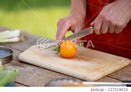Close up of man holding sharp knife, slicing juicy red tomato. Preparing vegetables for an outdoor barbecue. 117319018