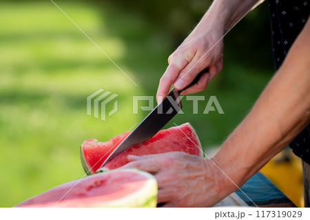 Close up of man holding sharp knife, slicing juicy red watermelon. Preparing food for an outdoor barbecue. 117319029