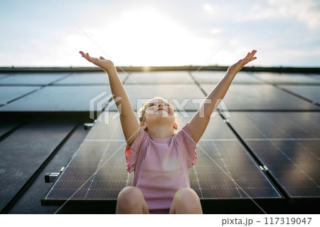 Cute girl on roof with solar panels, standing with open arms, hands up in the air. Rooftop solar or photovoltaic system. Sustainable future for next generation. Cute girl on roof with solar panels, standing with open arms, hands up in the air. Rooftop solar or photovoltaic system. Sustainable future for next generation. 117319047
