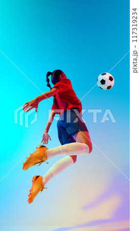 Soccer player leaps gracefully, arms outstretched, prepare to strike ball in neon light against colorful gradient background. 117319234