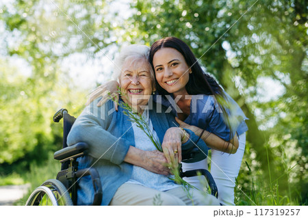 Female caregiver and senior woman in wheelchair. Nurse and elderly woman enjoying outdoor stroll during warm day. Female caregiver and senior woman in wheelchair. Nurse and elderly woman enjoying outdoor stroll during warm day. 117319257