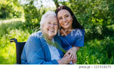Female caregiver and senior woman in wheelchair. Nurse and elderly woman enjoying outdoor stroll during warm day. 117319275