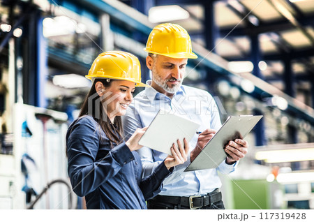 Two project managers standing in modern industrial factory, looking document in clipboard. Manufacturing facility with robotics, robotic arms and automation Two project managers standing in modern industrial factory, looking document in clipboard. Manufacturing facility with robotics, robotic arms and automation 117319428
