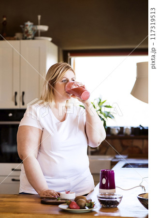 Overweight woman in the kitchen, drinking fresh smoothie. Choosing healthy food, making smoothie at home. 117319513