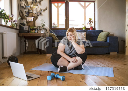 Online fitness training for overweight woman exercising at home, looking at laptop. Workout in living room. 117319538