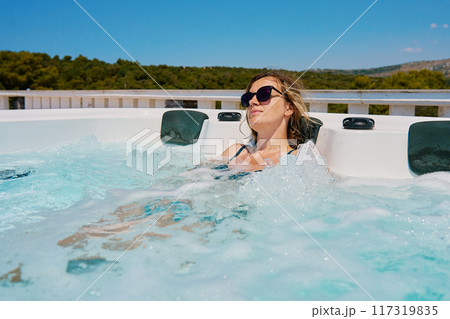 Woman wearing sunglasses and swimsuit relaxing in outdoor hot tub with bubbling water and enjoying sunny day. Female resting in outdoor jacuzzi with beautiful natural landscape 117319835