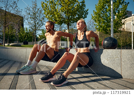 Sportive people, man and woman training outdoors in early summer morning, doing reverse push ups exercises, workout out in public park 117319921