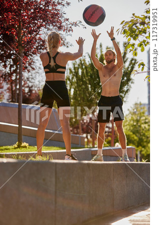Sportive man and woman enjoying intense outdoor workout, training with medicine ball outdoors in early sunny morning. 117319951