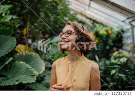 Beautiful woman standing in botanical garden, in the middle of lush green foliage with headphones on head, listening music. Beautiful woman standing in botanical garden, in the middle of lush green foliage with headphones on head, listening music. 117320717