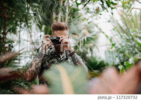 Handsome man with tattoos standing in botanical garden, in the middle of lush green foliage, holding camera and taking photography. Handsome man with tattoos standing in botanical garden, in the middle of lush green foliage, holding camera and taking photography. 117320732