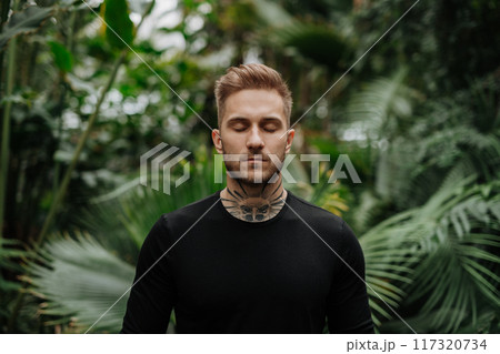 Handsome man with tattoo standing in botanical garden, in the middle of lush green foliage, looking at camera. Handsome man with tattoo standing in botanical garden, in the middle of lush green foliage, looking at camera. 117320734