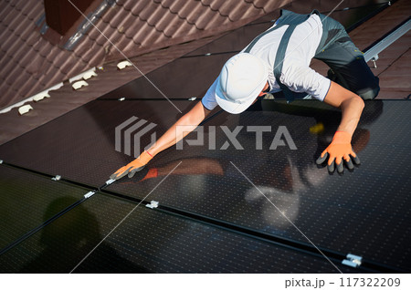 Worker building solar panel system on rooftop of house. Man engineer in helmet installing photovoltaic solar module outdoors. Alternative, green and renewable energy generation concept. Worker building solar panel system on rooftop of house. Man engineer in helmet installing photovoltaic solar module outdoors. Alternative, green and renewable energy generation concept. 117322209