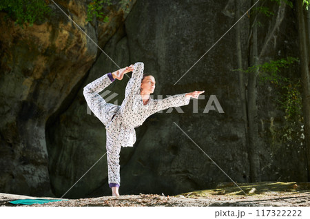 Woman practicing yoga outdoors in forest. Barefoot female on yoga mat surrounded by trees and large rocks, which suggests peaceful, natural environment ideal for meditation or yoga practice. Woman practicing yoga outdoors in forest. Barefoot female on yoga mat surrounded by trees and large rocks, which suggests peaceful, natural environment ideal for meditation or yoga practice. 117322222