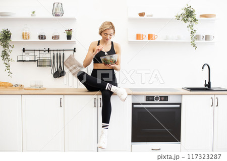Woman in athletic wear eating healthy salad in modern kitchen 117323287