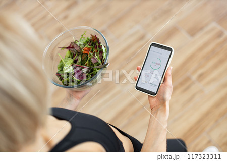 Woman tracking calories on smartphone while eating healthy salad Woman tracking calories on smartphone while eating healthy salad 117323311