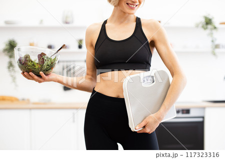 Fit woman holding salad bowl and bathroom scale in kitchen 117323316