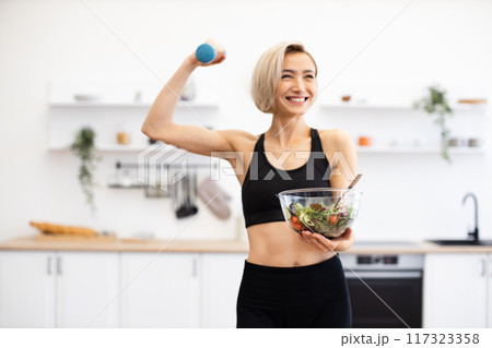 Fit woman holding salad and weight in modern kitchen 117323358