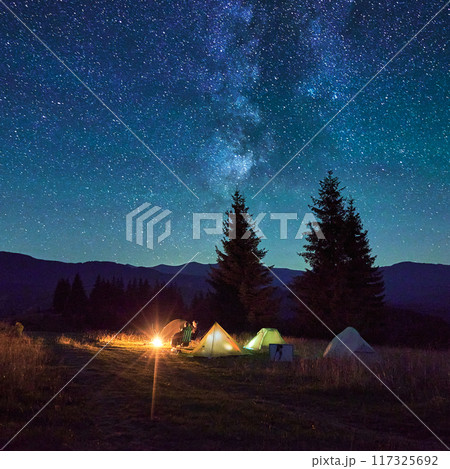 Two people tourists relax in chairs by campfire, with illuminated tent nearby, under breathtaking starry sky. Milky Way stretches above, creating magical and serene atmosphere in mountain wilderness. Two people tourists relax in chairs by campfire, with illuminated tent nearby, under breathtaking starry sky. Milky Way stretches above, creating magical and serene atmosphere in mountain wilderness. 117325692