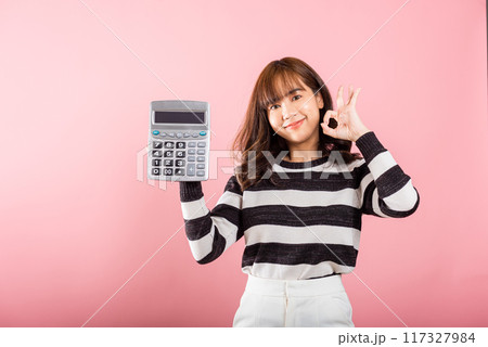 Tax day concept. Woman confident smiling holding electronic calculator and show OK gesture, excited happy Asian female studio shot isolated on pink background, Account and finance counting income Tax day concept. Woman confident smiling holding electronic calculator and show OK gesture, excited happy Asian female studio shot isolated on pink background, Account and finance counting income 117327984