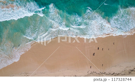 Aerial view of turquoise waves gently crashing onto a sandy beach. The oceans clear water, mixed with white foam, creates a tranquil 117328744