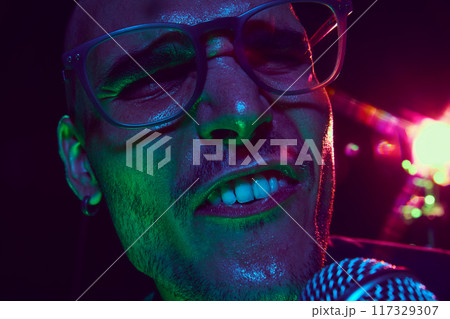 Vibrant lights illuminate singer's face, his open mouth and intense expression capturing raw energy of live performance. Vibrant lights illuminate singer's face, his open mouth and intense expression capturing raw energy of live performance. 117329307