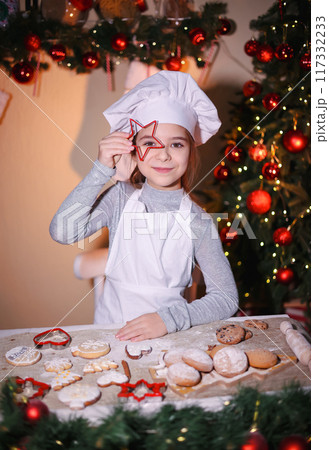 Cheerful girl in chef's hat shows looking through baking pan of cookies on Christmas background 117332233