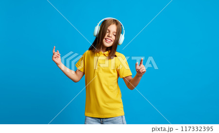 Young girl in yellow polo shirt enjoying music in headphones, smiling against blue studio background. Entertainment and leisure 117332395
