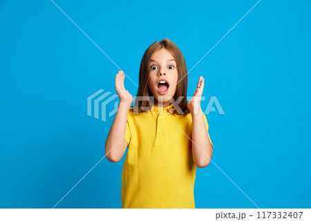 Portrait of young emotional girl in yellow polo shirt expressing shock and excitement against blue studio background. Surprise Portrait of young emotional girl in yellow polo shirt expressing shock and excitement against blue studio background. Surprise 117332407