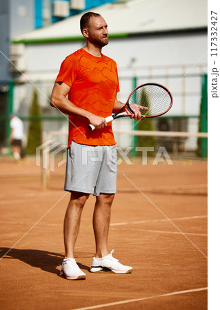 Full-length image of man, tennis player in orange t-shirt standing with racket on outdoor clay court before of after intense match. Full-length image of man, tennis player in orange t-shirt standing with racket on outdoor clay court before of after intense match. 117332427