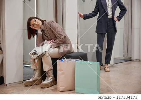 Bored female feeling tired while her friend fitting new clothes attire Bored female feeling tired while her friend fitting new clothes attire 117334173