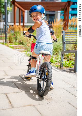 Little girl rides a two-wheeled bicycle Ii the courtyard of apartment buildings in summer, Europe Little girl rides a two-wheeled bicycle Ii the courtyard of apartment buildings in summer, Europe 117334343