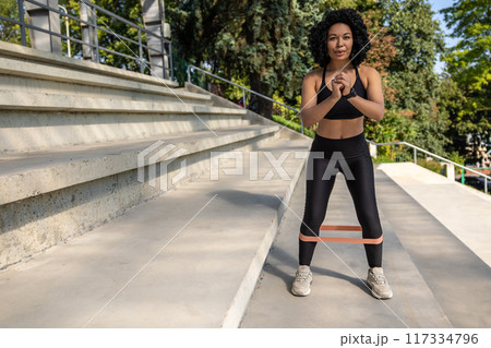 Fit woman in black sportswear exercising outdoors with a power band on her legs 117334796