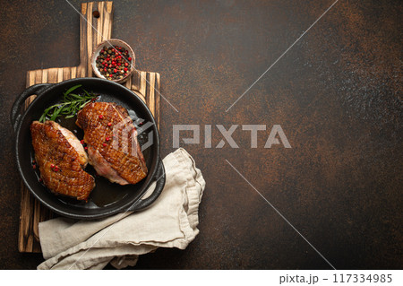 Two roasted duck breast fillets with crispy skin, with pepper and rosemary, top view in black cast iron pan with knife, dark brown concrete rustic background, space for text Two roasted duck breast fillets with crispy skin, with pepper and rosemary, top view in black cast iron pan with knife, dark brown concrete rustic background, space for text 117334985