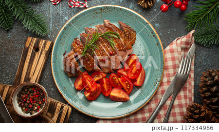 Festive Christmas dinner with sliced roasted duck breast fillet with golden crispy skin and tomato salad, Xmas decorations on table with fir tree, cones, red berries, blue rustic concrete background. 117334986