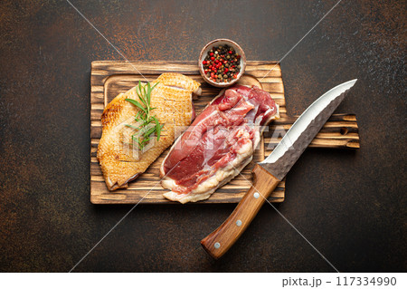 Two raw uncooked duck breast fillets with skin, seasoned with salt, pepper, rosemary top view on wooden cutting board with knife, dark brown concrete rustic background 117334990