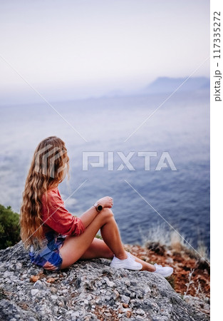 Woman sitting on a rock by the sea at sunset in a coastal location 117335272