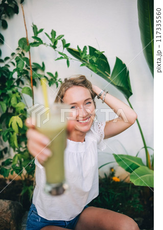 Young woman enjoys a refreshing green drink surrounded by tropical plants on a sunny day 117335560