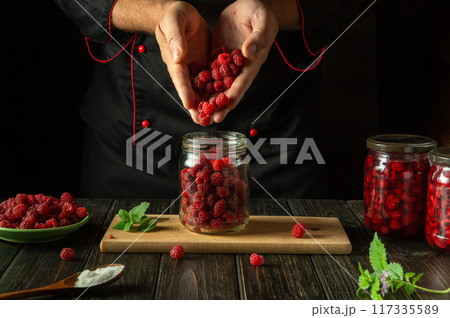 The cook prepares jam with Rubus idaeus. The concept of making sweet raspberry jam on the kitchen table. Filling the jar with ripe berries in the hands of the chef The cook prepares jam with Rubus idaeus. The concept of making sweet raspberry jam on the kitchen table. Filling the jar with ripe berries in the hands of the chef 117335589